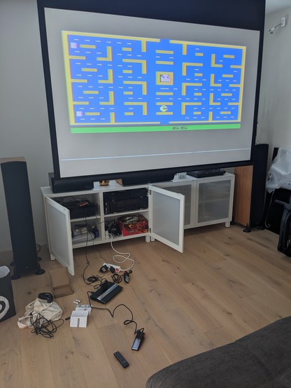 Game console with controllers and cables on the floor and PacMan on the projector image above.