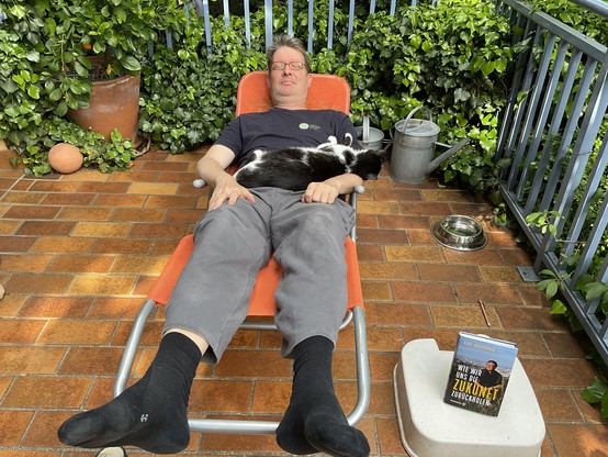 Kury liegt schlafend mit hochgelagerten Füßen auf einer Sonnenliege. Auf seinem Buch liegt ein schwarz-weißer Kater. Im Vordergrund das Buch: „Wie wir uns die Zukunft zurückholen“ von Rudi Anschober