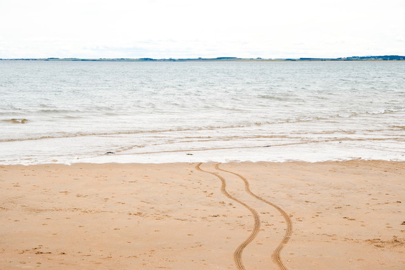 Ein Meeresstrand aus Sand. Aus dem Wasser führen direkt zwei Reifenspuren in Richtung Kamera.