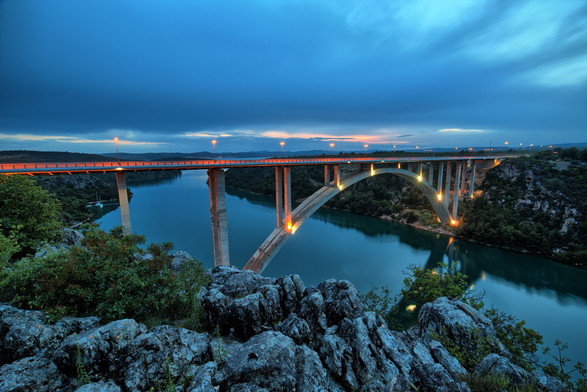 Eine Brücke, die stellenweise beleuchtet ist führt von einem Ufer zum anderen. Es ist eine Langzeitaufnahme und man sieht die langezogenen Lichter der Autos. Im Vordergrund ist Felsen. Die Brücke führt über einen Fluss. Der Himmel ist bewölkt.