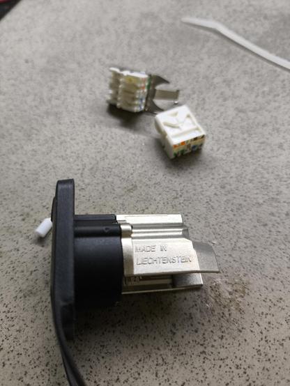 Auf einem Tisch ist ein offener Netzwerk Stecker zu sehen. Darauf geschrieben steht "Made in Liechtenstein". Dahinter weitere Bauteile.