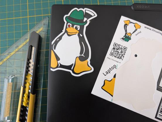 A Lenovo X13 2-in-1 notebook upper left corner where a Linuxtage Steirertux sticker is put above the Thinkpad logo. One eye of the Steirertux is cut out so that the Lenovo status LED is visible from behind, resembling the red eye of a T1000 from the Terminator movies.