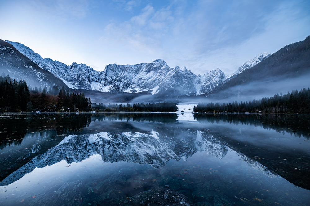 Vorne ein See, dahinter Ufer mit Nadelbäumen. Im Anschluss erhebt sich ein großes Gebirgsmassiv. Es herrscht blaue Stunde und über dem See sind leichte Nebelschwaden.