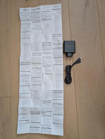 Die Bedienungsanleitung eines USB Netzteils neben dem Netzteil. Man sieht nur eine der beiden Seiten vom Manual. Es ist voll mit sehr kleiner Schrift.