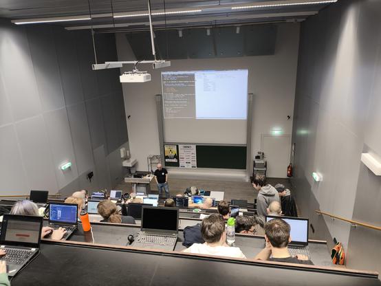 Ein Universitätshörsaal mit mehreren Sitz-Reihen, einige Personen sitzen mit Laptops vor sich. Vorne steht der Vortragende, hinter ihm ist die Präsentation an der Wand zu sehen. 