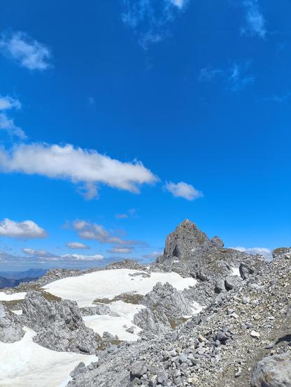 Ein spitzer felsiger Berg in der Bildmitte. Darunter eine Ebene die teilweise schneebedeckt ist. Blauer Himmel.