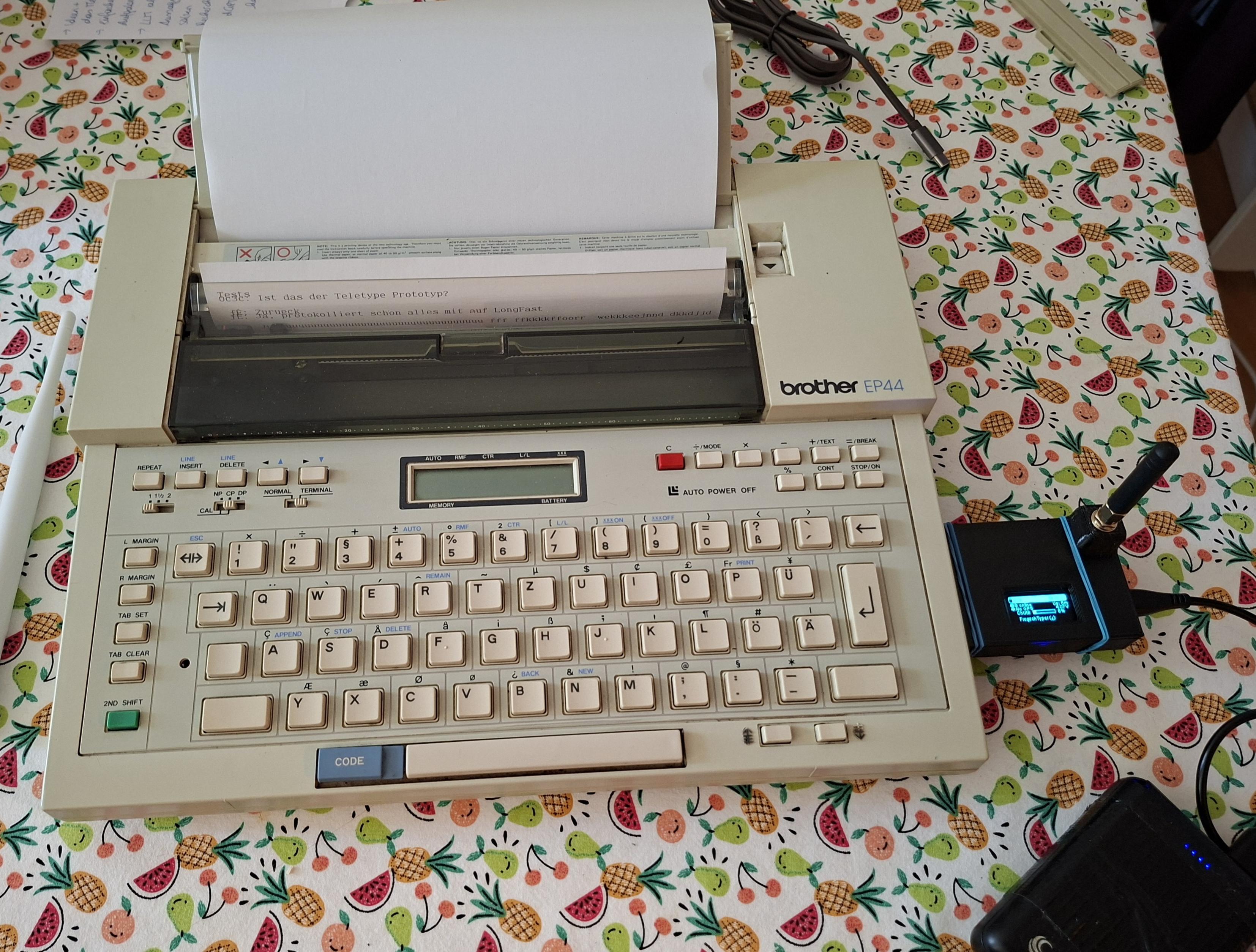 A Brother EP44 typewriter on a table with a flower cloth. Attached to the side a Meshtastic node for forwarding messages to and from the mesh network