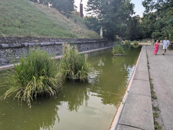 Schanzgraben with greenish colored water, plants, a hill, people in Stadtpark Graz