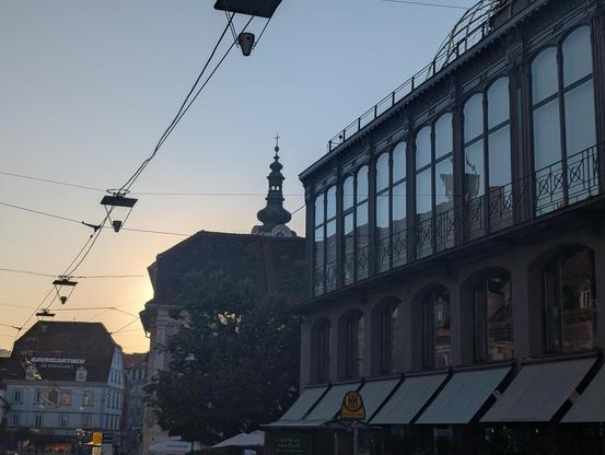 great colors in the sky with darker buildings in the foreground, things visible in glass windows, a church tower and some tramway cables in the air